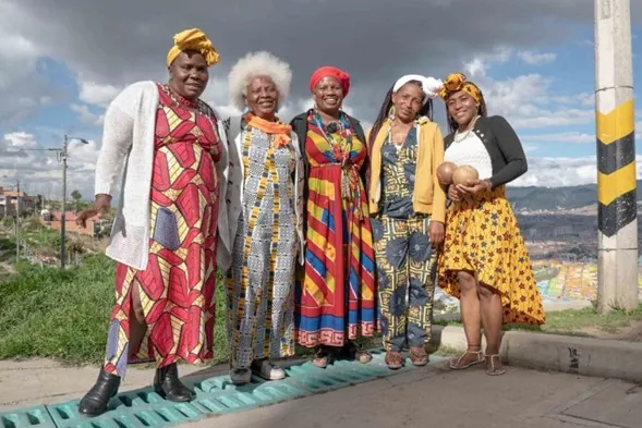 Cinco mujeres afrodescendientes con turbantes y vestidos tradicionales de colores, de pie en una calle de montaña