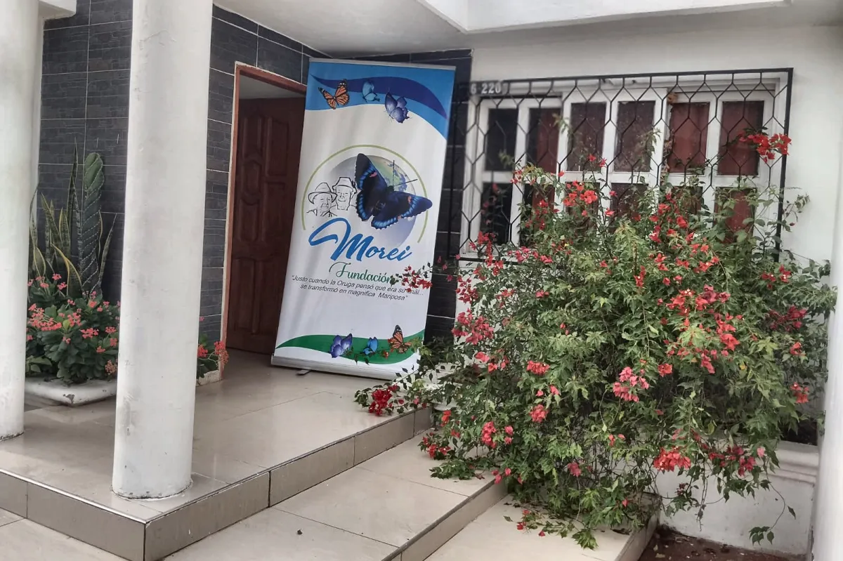 Entrada de la sede en Campo de la Cruz: columnas blancas, el pendón de la Fundación Morei y una buganvilia florecida junto a la ventana